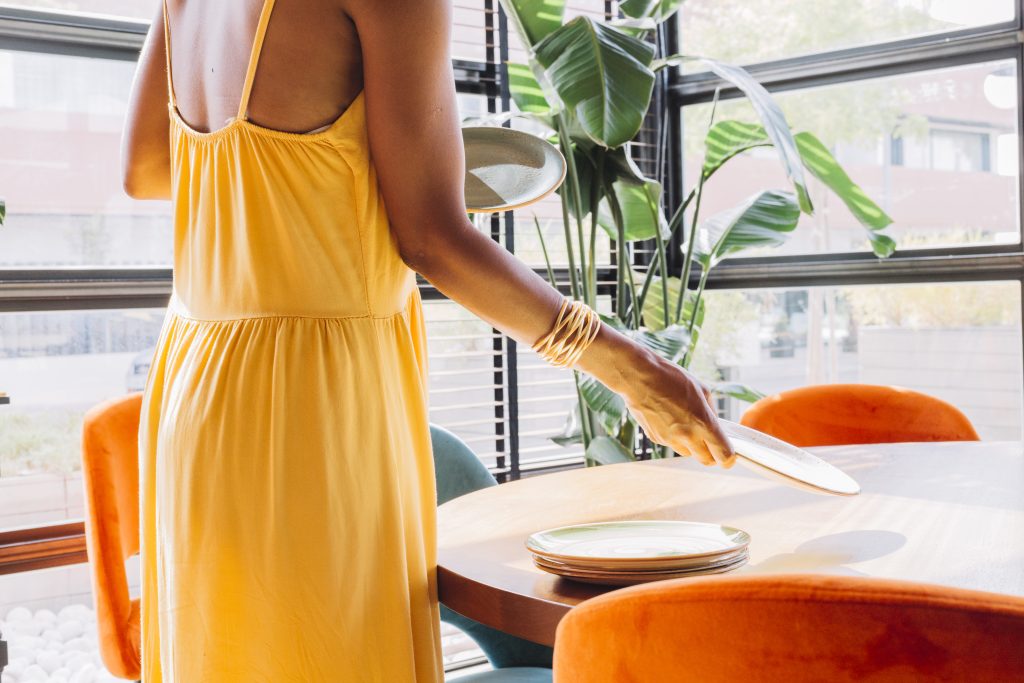 Uma mulher vestida de amarelo, pondo os pratos na mesa de sua varanda gourmet.